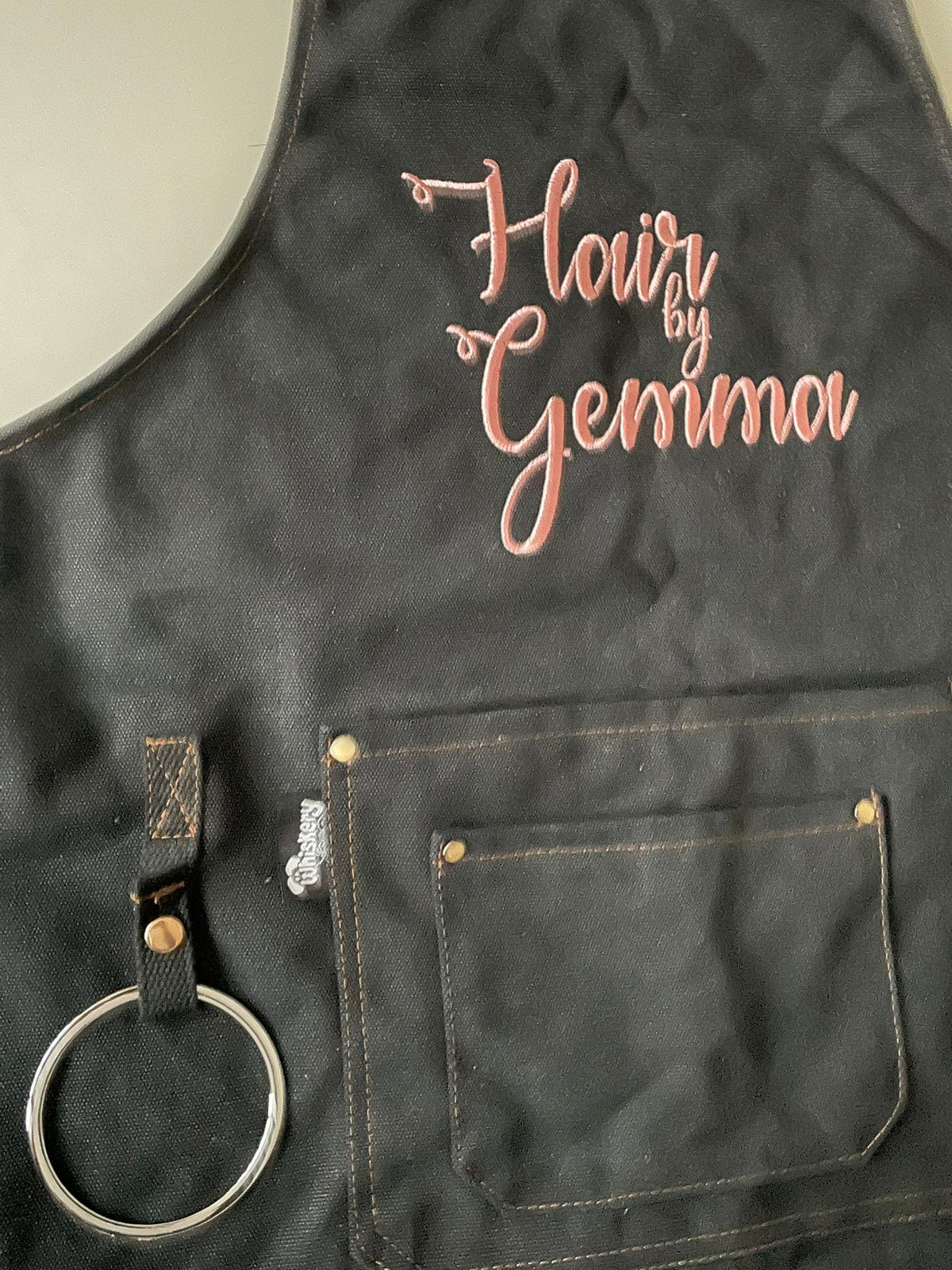 Black canvas apron embroidered with Hair by Gemma in pink script thread — personalised salon apron | Whiskery Sour